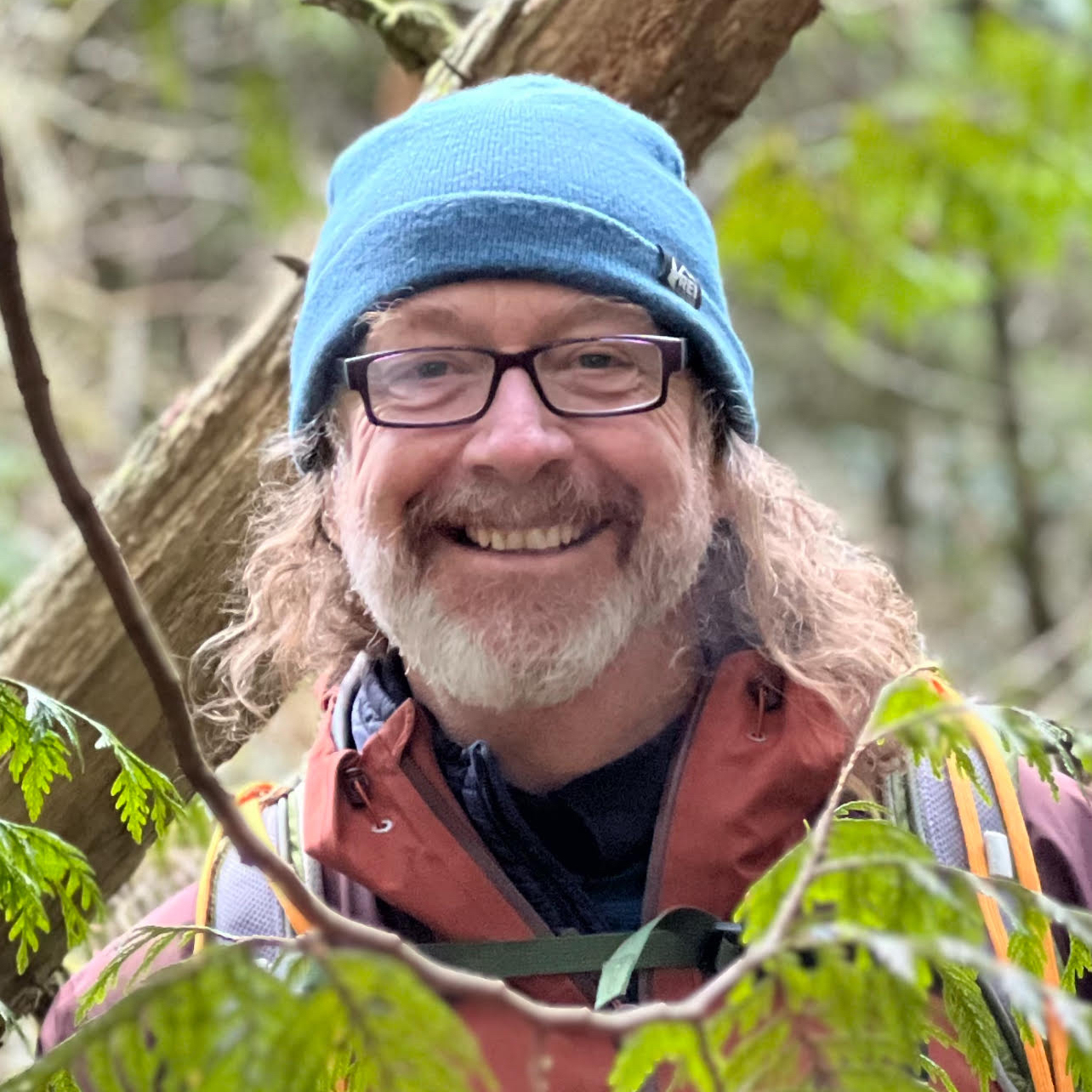 Dr Sherwood in a blue hat and burgundy rain jacket smiles in the forest, among the trees.
